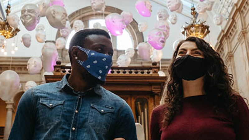 Two people wearing face masks walking in Kelvingrove Art Gallery. In the background there is a floating heads installation which shows heads suspended from the ceiling with lots of different facial expressions. 