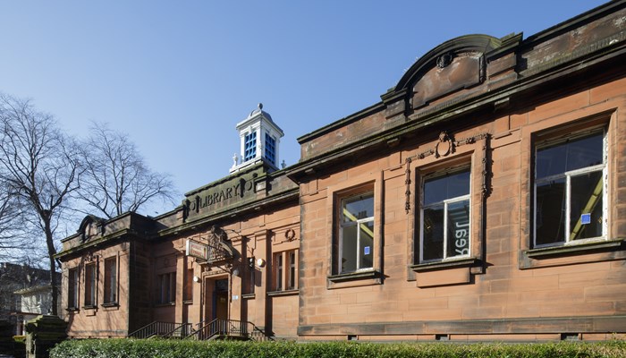 An exterior close-up image of Langside Library which is a red sandstone building built in 1915. There is a 'Library' sign carved into the building above the doorway and also a projecting 'Public Library' sign. There are 3 large windows at each side of the entrance. There are hedges surrounding the front of the building. This photograph has been taken on a sunny day in winter - you can see a bare tree in the background. 