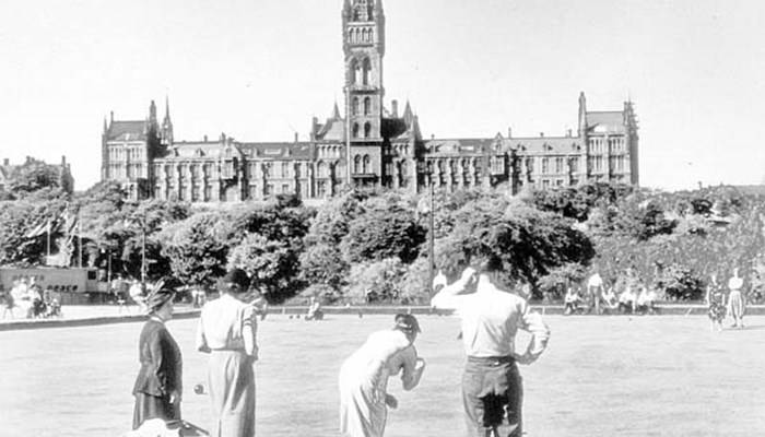 A black and white photo of a group of people playing lawn bowls with Glasgow University in the distance.