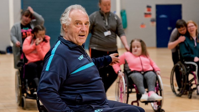 Man in a wheelchair, in a sports hall setting and pointing to a group of people in the background, 3 of the people are also in wheelchairs.