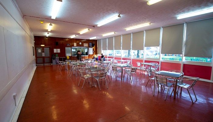 large room with multiple tables are chairs places around the room. the floor is dark orange and the walls are white. there are windows lining the right hand side of the room