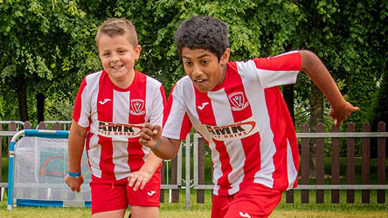 Two children running towards the camera playing football. They both have a red and white strip on and are laughing. The game is taking place outdoors.