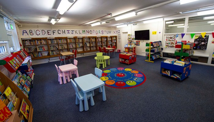 A view of the spacious children's area. There is an alphabet above a row of bookshelves, colourful tables and chairs, books and a rug with faces in different colours.
