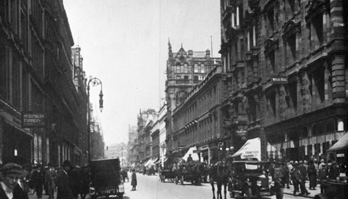 A black and white photo of a past Buchanan Street in Glasgow, with tall sandstone buildings on both sides, horse and carriages, parked cars and many people walking on the pavements.