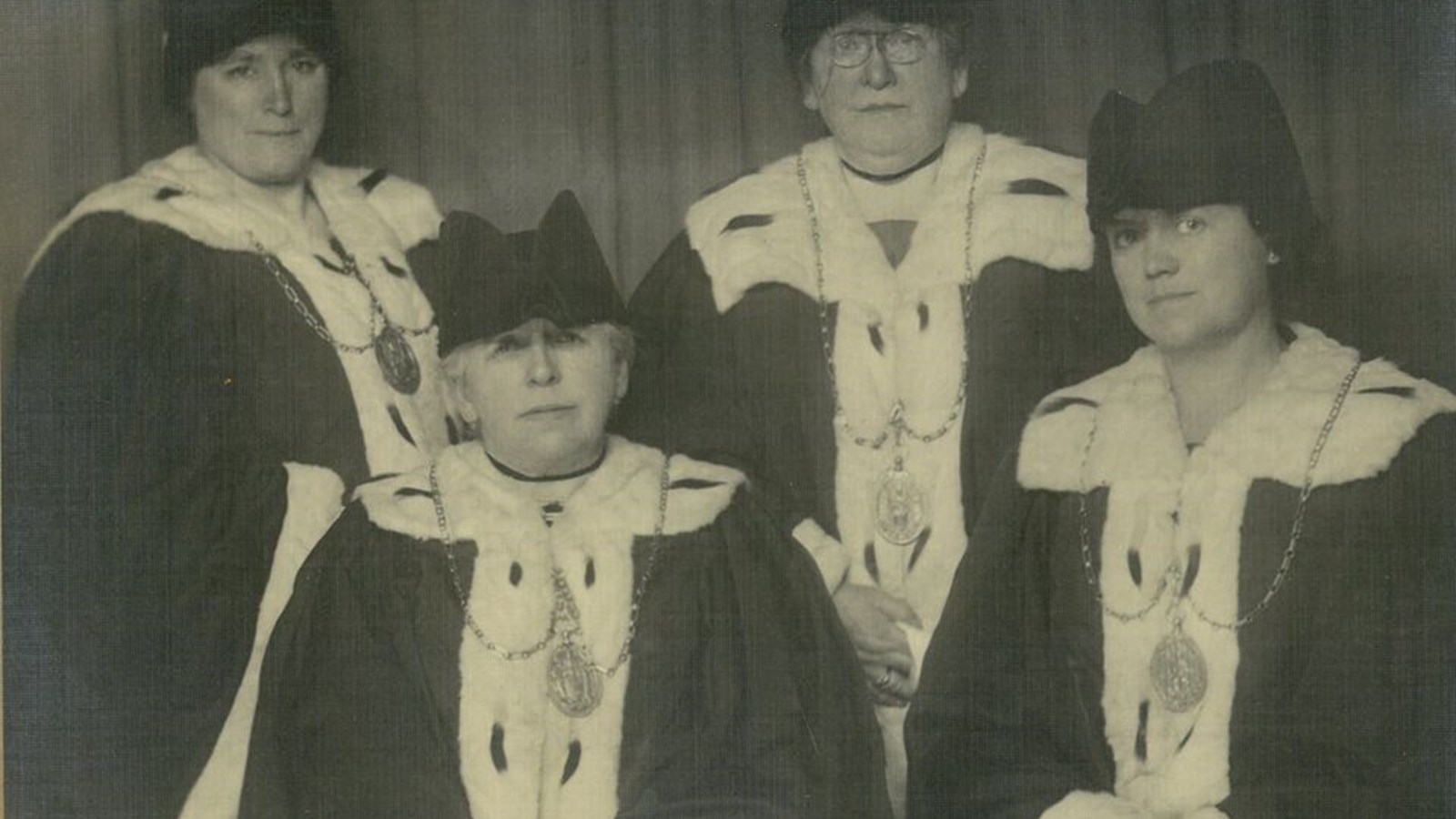 Black and white photo of Mary barbour sitting end left with three other people wearing their ceremonial robes which includes a black hat, white fluffy collar, lapels and cuffs and a large medallion.