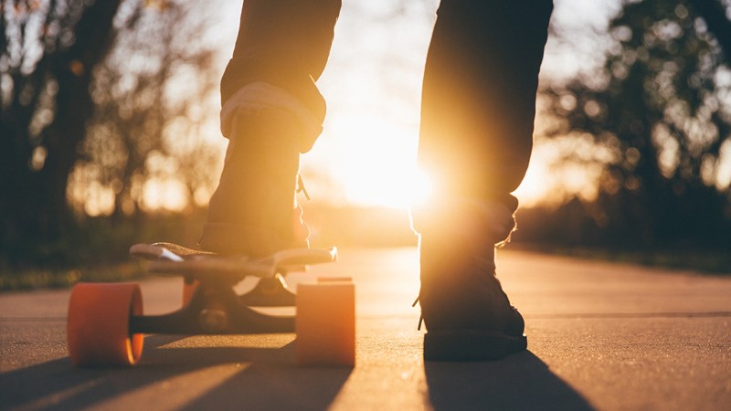 The silhouette of a young person standing with their left foot on a skateboard. The sun is low in the sky in the background.