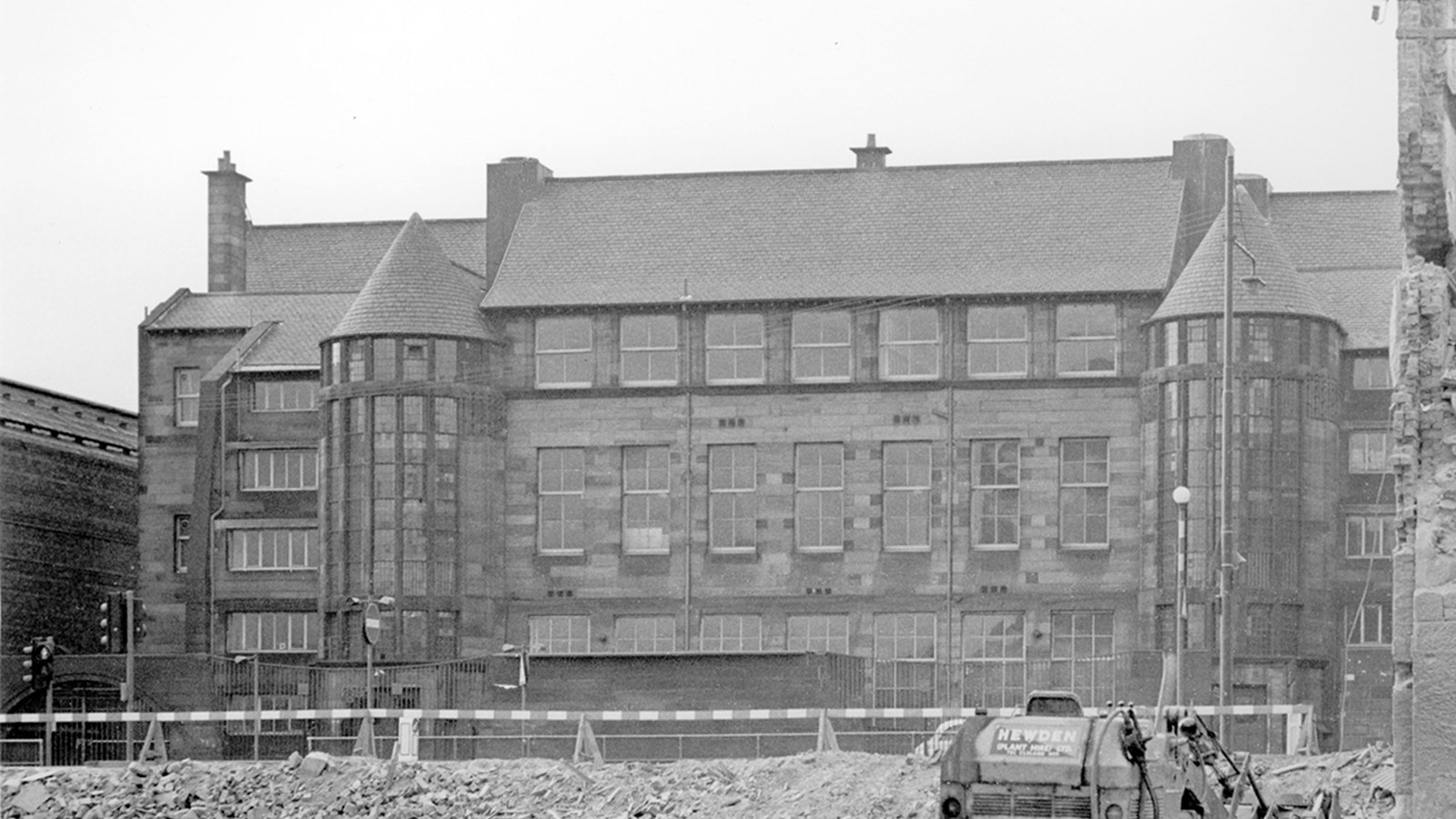A school board building in black and white photography receiving development work outside, with a small digger and barriers in the foreground.