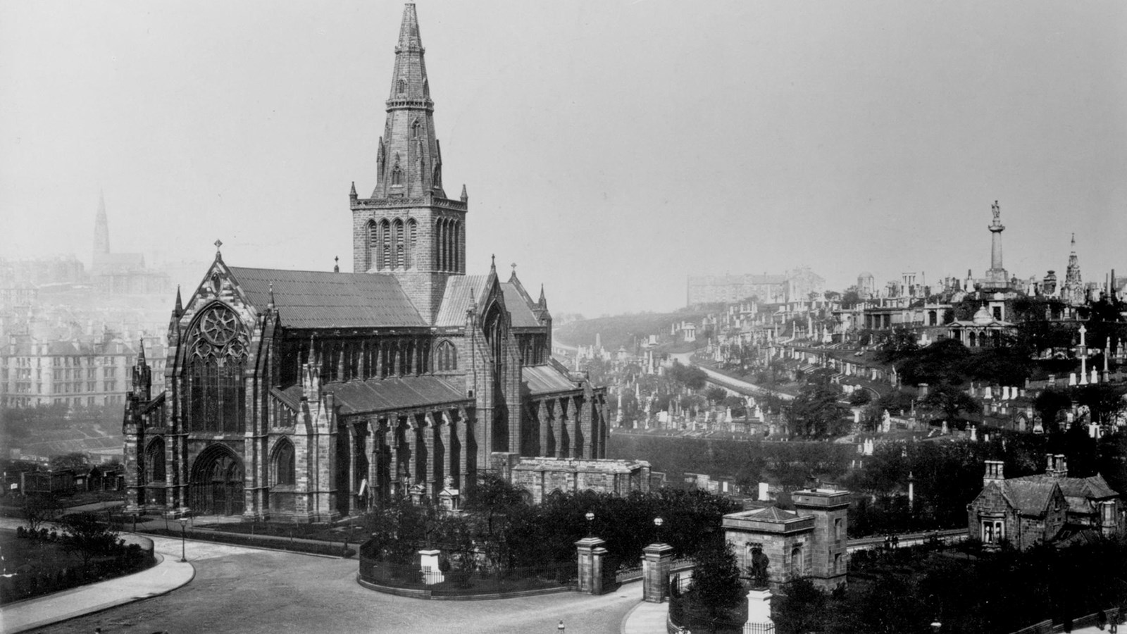 A black and white photo of The Glasgow Necropolis with the city in the background. People are sitting on benches on the streets and a horse and carriage is bypassing.