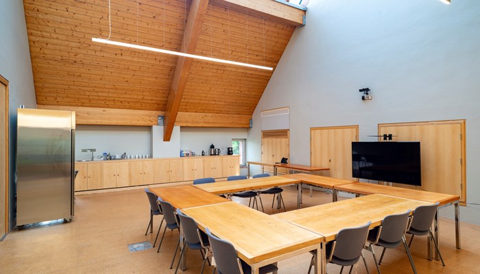 large meeting space with two large tables with chairs round them. there is a kitchen area and a fridge on one side of the room