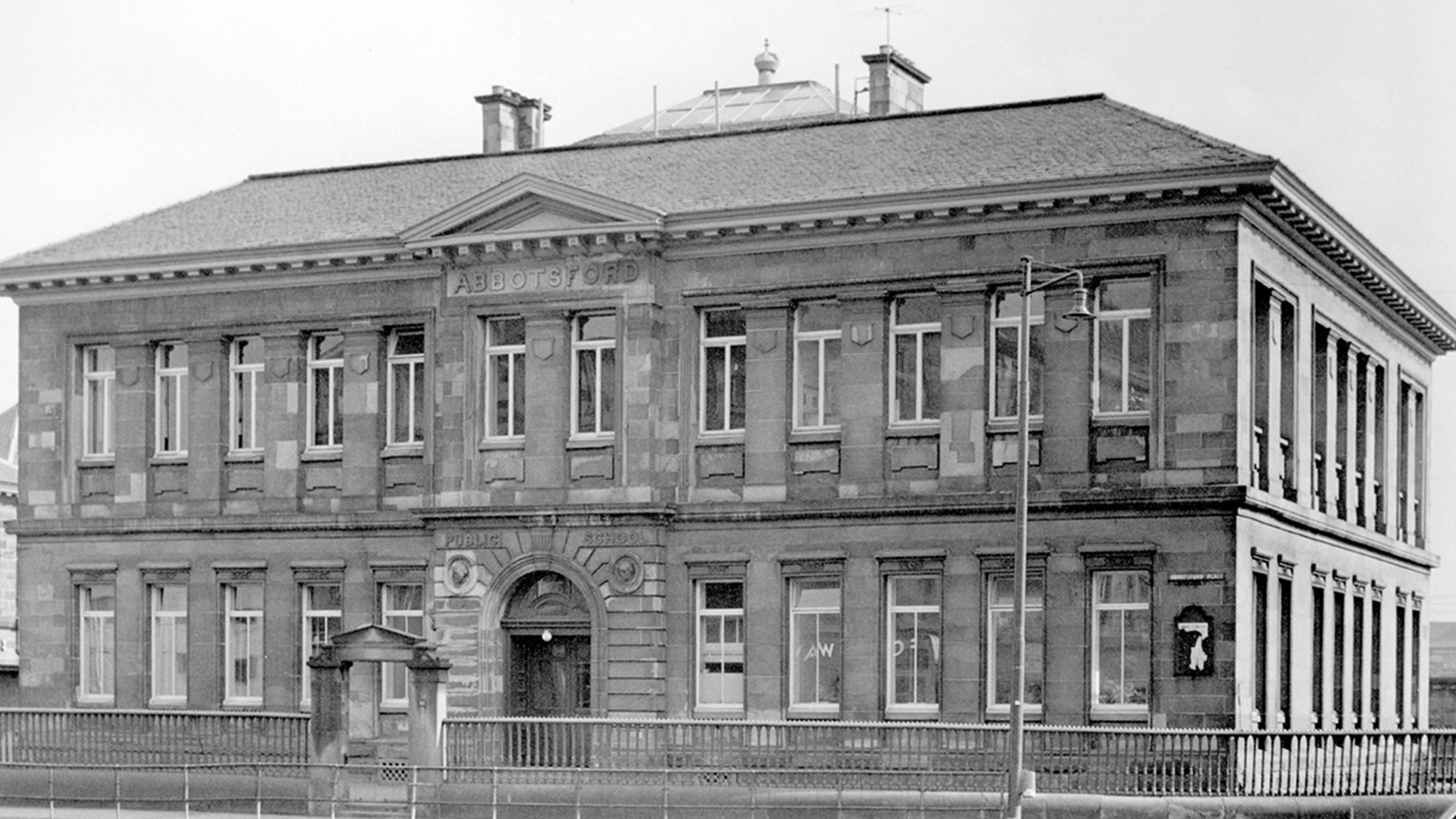 A black and white photo of three story Abbotsford School (1879) with white window frames and an arch entry via the main gates.