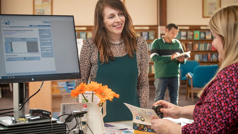 A person standing at a Glasgow library desk chatting to a staff member and getting books stamped. There is another person in the background reading a book and there are lots of book shelves. There is a library computer and a vase of orange flowers in the foreground.