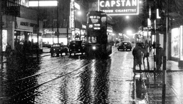 A black and white photo of a scene of Glasgow at night in the rain, with lit up signs, trams, cars and pedestrians.