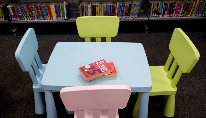 Children's table and chairs at Anniesland Library. The table and 1 chair is light blue. There are two yellow chairs and one pink chair. In the background there is a bookshelf filled with books.