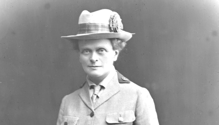 A black and white photo of Elsie Maud in a light coloured uniform, including a tie and a hat, grasping a pair of gloves in her left hand.
