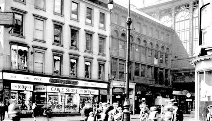 Black and white photo showing busy street with store fronts and groups of people crossing the road