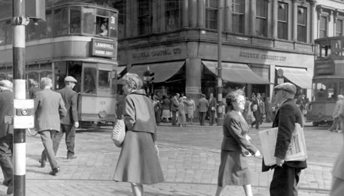 A black and white photo of a busy street with trams, many pedestrians, high sandstone shop buildings and traffic lights.