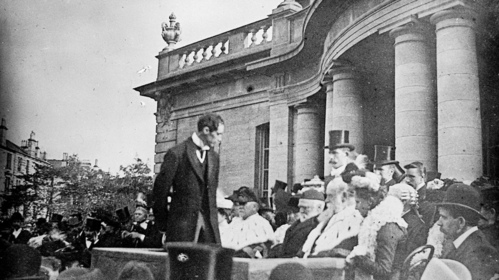 A black and white photograph of a crowd of people standing outside a historical building dressed in suites and dresses and many wearing hats including top hats.