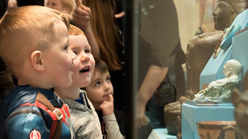 Three young children looking in awe at a display of Chinese figures in a glass case at the Burrell Collection