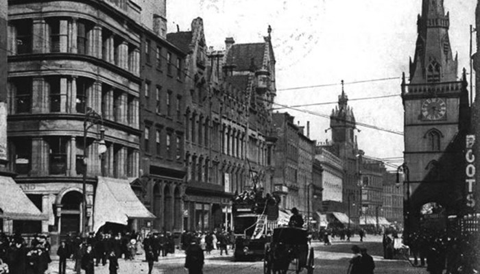 A black and white photo of a busy street with trams, horse and carriages, pedestrians, many sandstone tall shop buildings and a clock tower.