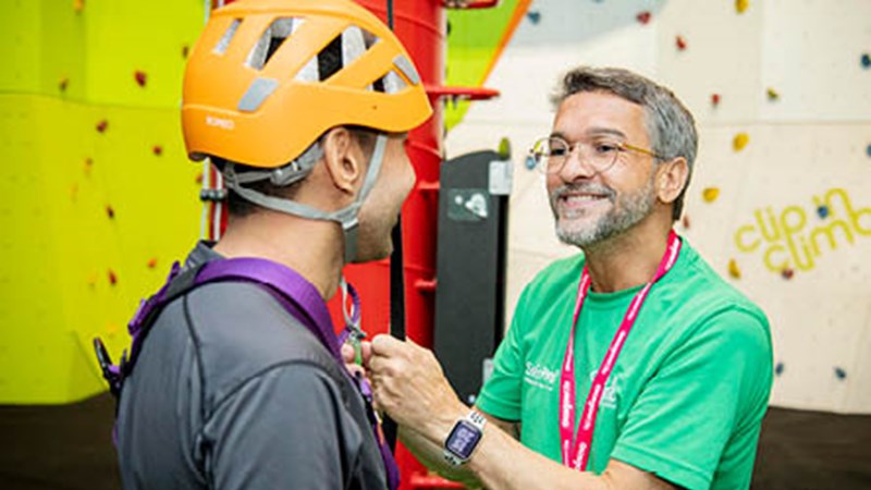 A staff member with a Glasgow Life lanyard smiling as they help to fit a harness to a person about to do clip and climb - climbing experience at Kelvin Hall. The other person has a helmet on and has their back to the camera.