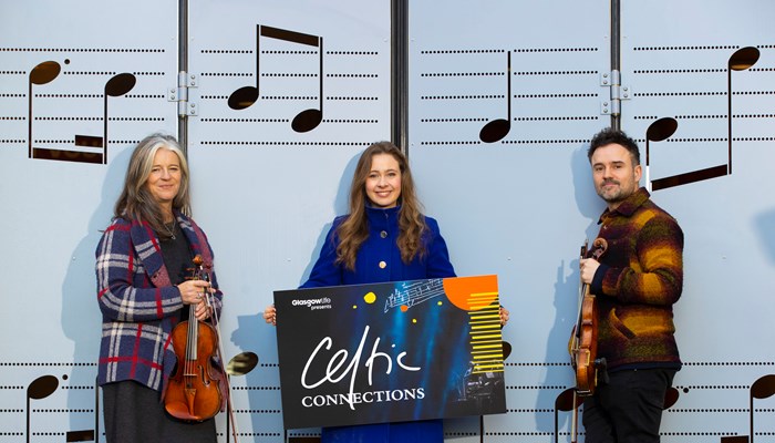Three musicians standing in front of large-format sheet music showing notes