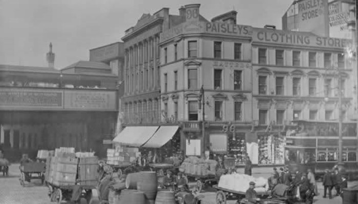 A black and white photo of a busy street in Glasgow city centre which has many horse and carriages carrying heavy loads of barrels and other materials. People are working in the street to get everything packed up. The street has large and tall buildings and a bridge in the background. 