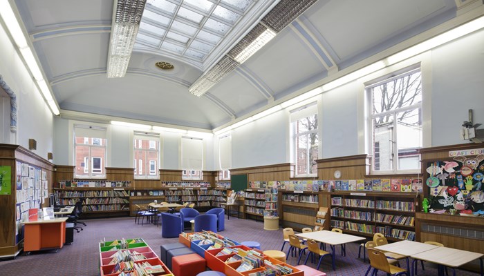 A large spacios childrens area which is long and rectangular. There are wooden bookshelves filled with colourful children's books along the perimeter of the room. There is a high curved ceiling with a long grid-colonial style skylight. There is wooden panelling around a third of the walls. The rest of the walls and ceiling are painted white. There is also colourful children's artwork on the walls, tables and chairs and colourful book storage boxes in the middle of the room. 