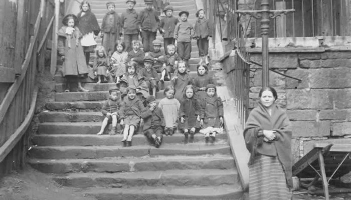 Black and white photograph of a group of children of all ages posing on steps. Older children are in the back rows standing and younger children are on front rows sitting. There is one child are the very back holding a baby and an adult at the front right looking at the camera.