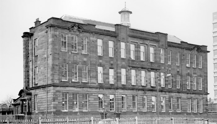 A black and white photograph of Sir John Maxwell School that is rectangular in shape with many rectangle shaped windows. A car is parked at the front of building and a high rise flat is in the background.