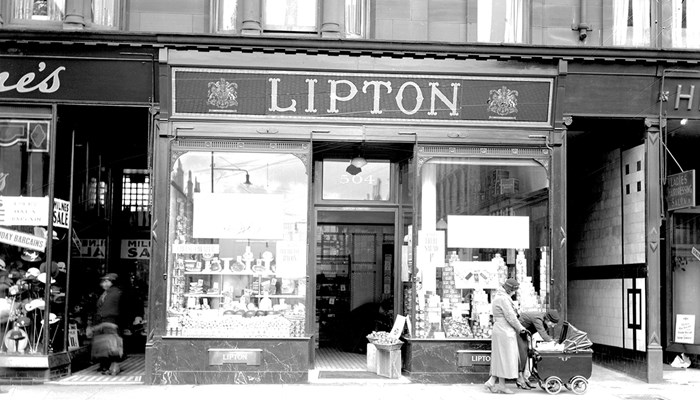A black and white photo of Lipton's shop front underneath tenement flats, with large glass windows showing their displays, with two people passing by pushing a baby in a pram.