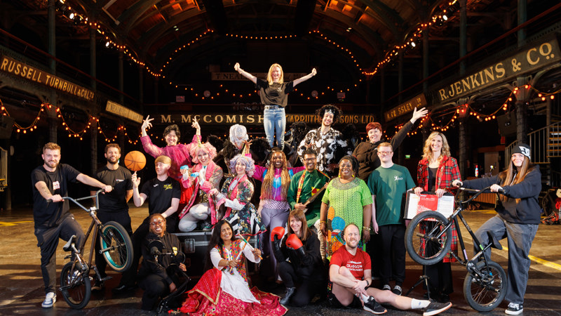 A diverse group of people pose together in a festive indoor setting, wearing colorful costumes and casual clothing, with props like bikes, musical instruments, sports gear, and boxing gloves, celebrating creativity and community.