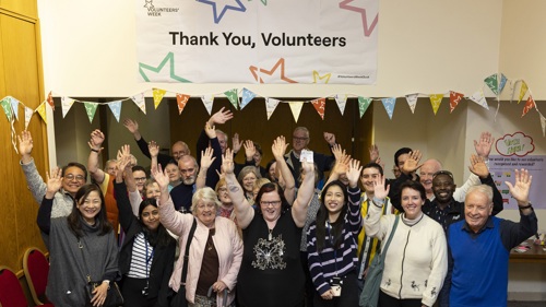 A large group of people at a celebration event in a wood panelled room under a Thank You Volunteers banner. They are smiling and have their hands in the air
