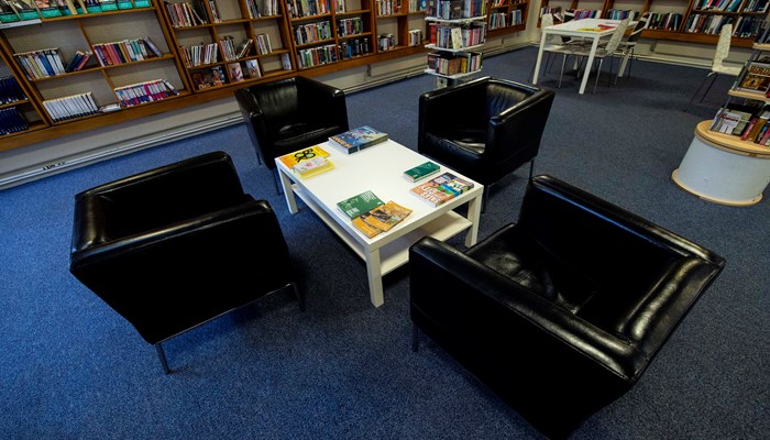 An inside view of Bailliston Library. The library has dark brown bookshelves and a blue carpet. There are 4 black chairs surrounding a white table.