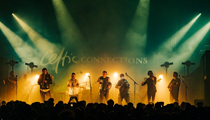 A six-piece band playing string instruments and drums on stage at Celtic Connections with spotlights on them.