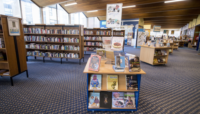 The main library area showing rows of wooden bookshelves, there are standalone bookshelves in the middle of the room. The ceiling is high and has wood pannelled.