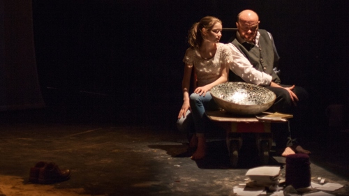 Two actors on stage with dark background and various props in front of them during Gaelic Arts at Tramway