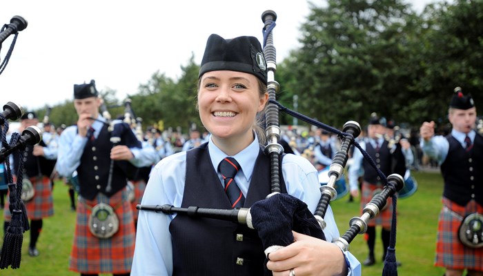 A bagpiper smiles to camera with fellow bandmate pipers out of focus in the background in an outdoor setting