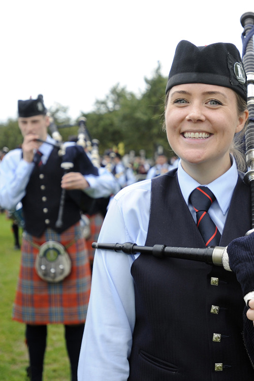A bagpiper smiles to camera with fellow bandmate pipers out of focus in the background in an outdoor setting
