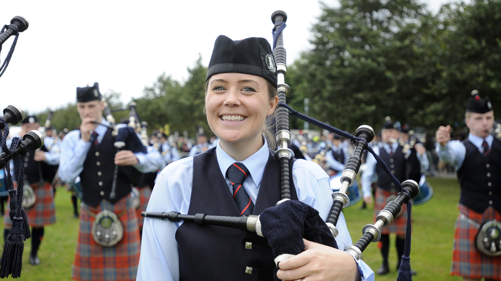 A bagpiper smiles to camera with fellow bandmate pipers out of focus in the background in an outdoor setting