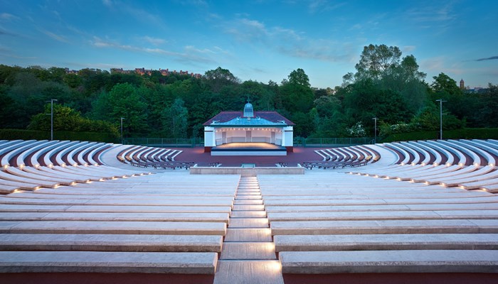 An empty Kelvingrove Bandstand with tall green trees in the background and curved concrete seating, pictured in evening light