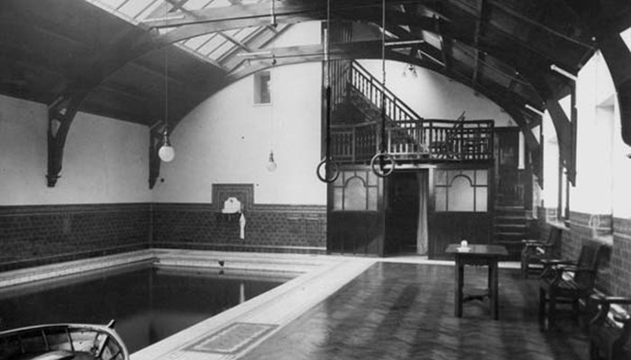 A black and white photo of the inside of an old gymnasium and swimming pool, with an empty rectangular pool and gymnastic rings hanging from the ceiling.