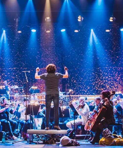 Conductor and live orchestra on stage under blue lights.