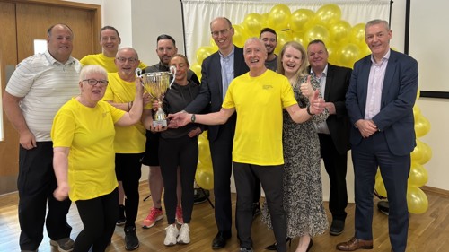 A large group of people gathered for a photograph in a sports hall to celebrate with a large silver trophy. Some of them are wearing the same yellow t-shirts and there is a yellow balloon arch behind them.