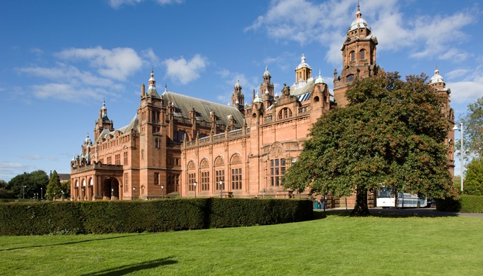 External view of the front of Kelvingrove Art Gallery & Museum with a bright blue sky in the background with a tree and grass lawn to the front of the building