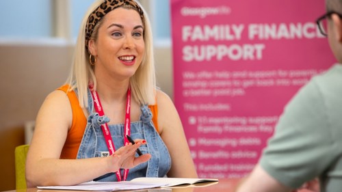 Two people are sitting across a table from each other in an office or library building. One is smiling and holding a pen in their hand with an open notebook on the table