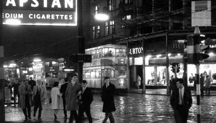 A black and white photo at night time of a street, the ground is wet from rain and people are walking on the pavement and crossing the road. Lights are omitted from shops, signs and trams.