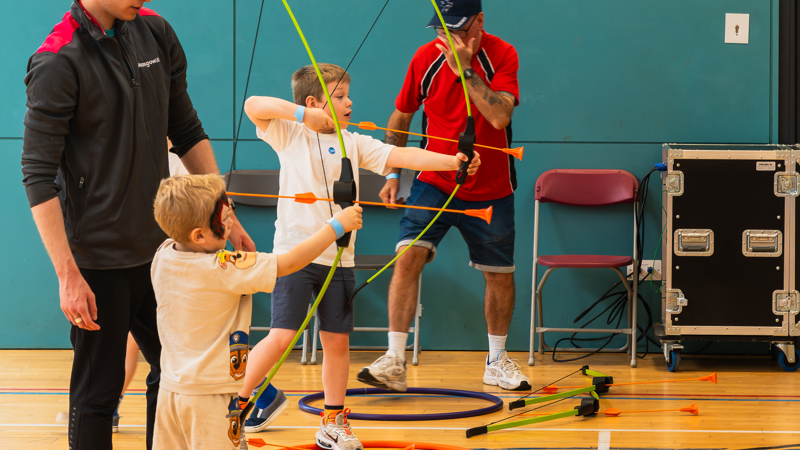 Two young boys are practicing archery indoors with guidance from two adult instructors, with bows drawn and arrows aimed at targets off-camera.