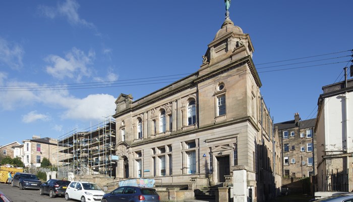 Exterior of Dennistound Library which is a 2-storey, blonde sandstone building with large windows and a dome with statue on top. This image was taken on a sunny day. There are 4 cars parked outside the library.