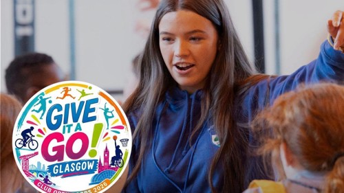 A sports coach with long hair is leading a session with young people in a sports hall