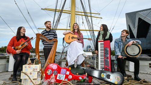 Five musicians performing on the deck of a tall ship surrounded by ropes, instrument cases and maritime rigging.